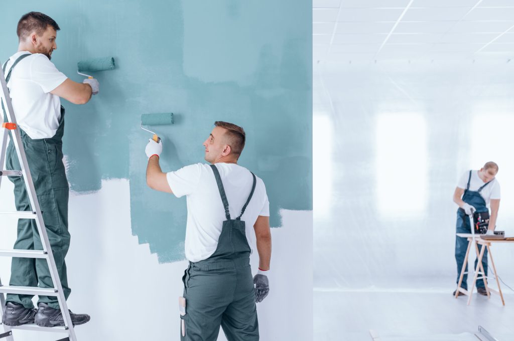 Men painting the wall blue using rollers in empty home interior